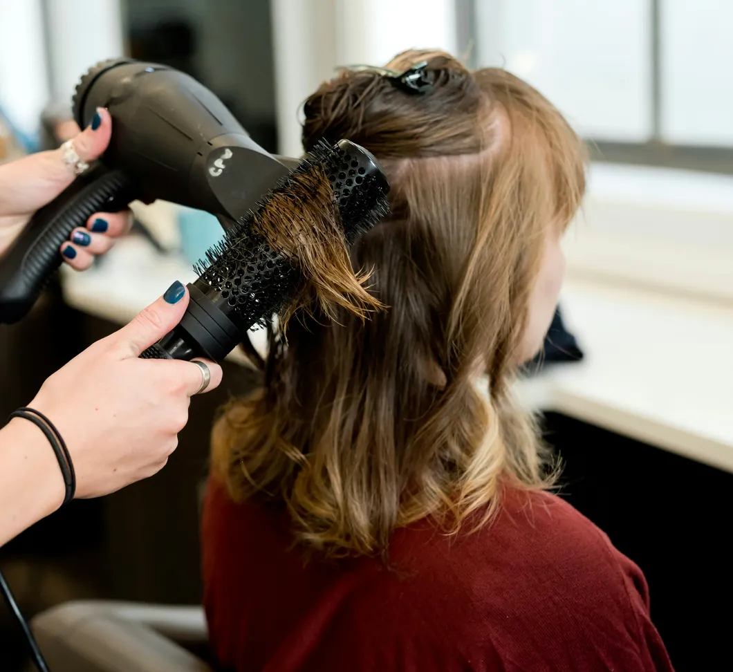 Coiffeuse en train de faire un brushing à une cliente vue de dos, dans un salon lumineux